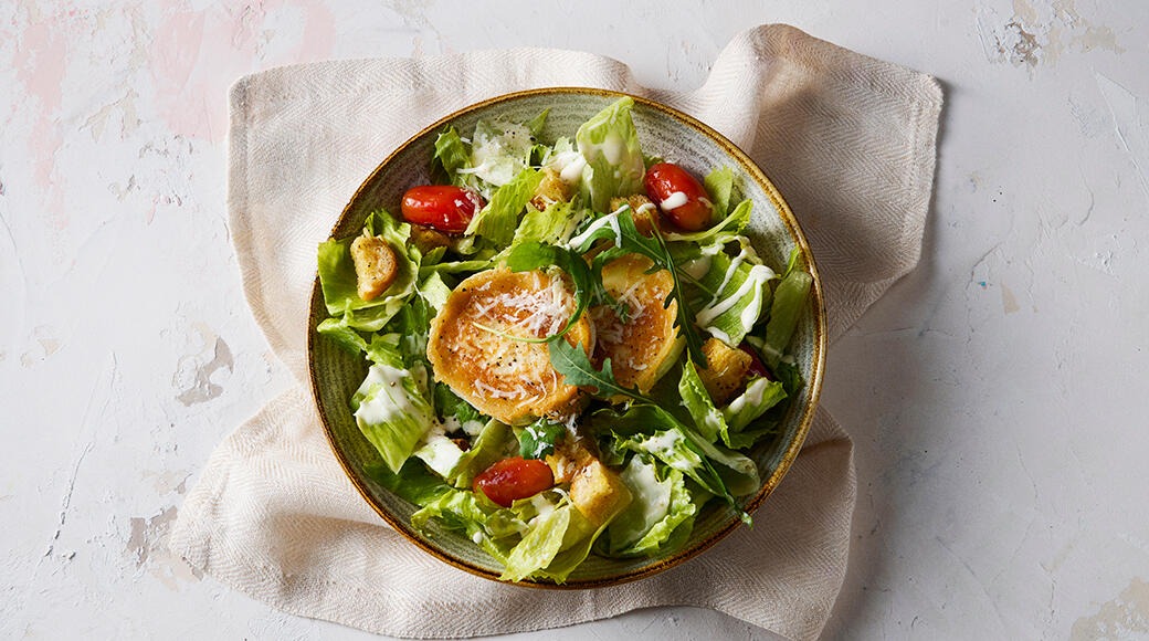 Bowl of salad with lettuce, rocket, cherry tomatoes, croutons, grilled cheese and dressing. The bowl sits on a cloth napkin.