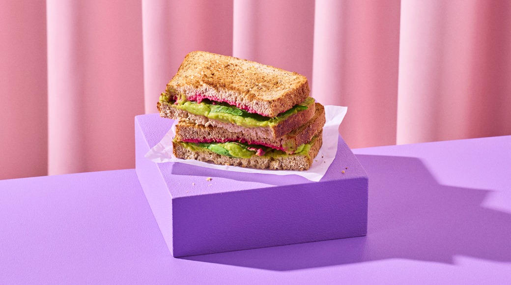 Paahdettu täysjyväleipä-voileipä, jossa avokadoa ja punaista levitettä, pinottuna violetin korokkeen päällä vaaleanpunaista verhoa vasten.