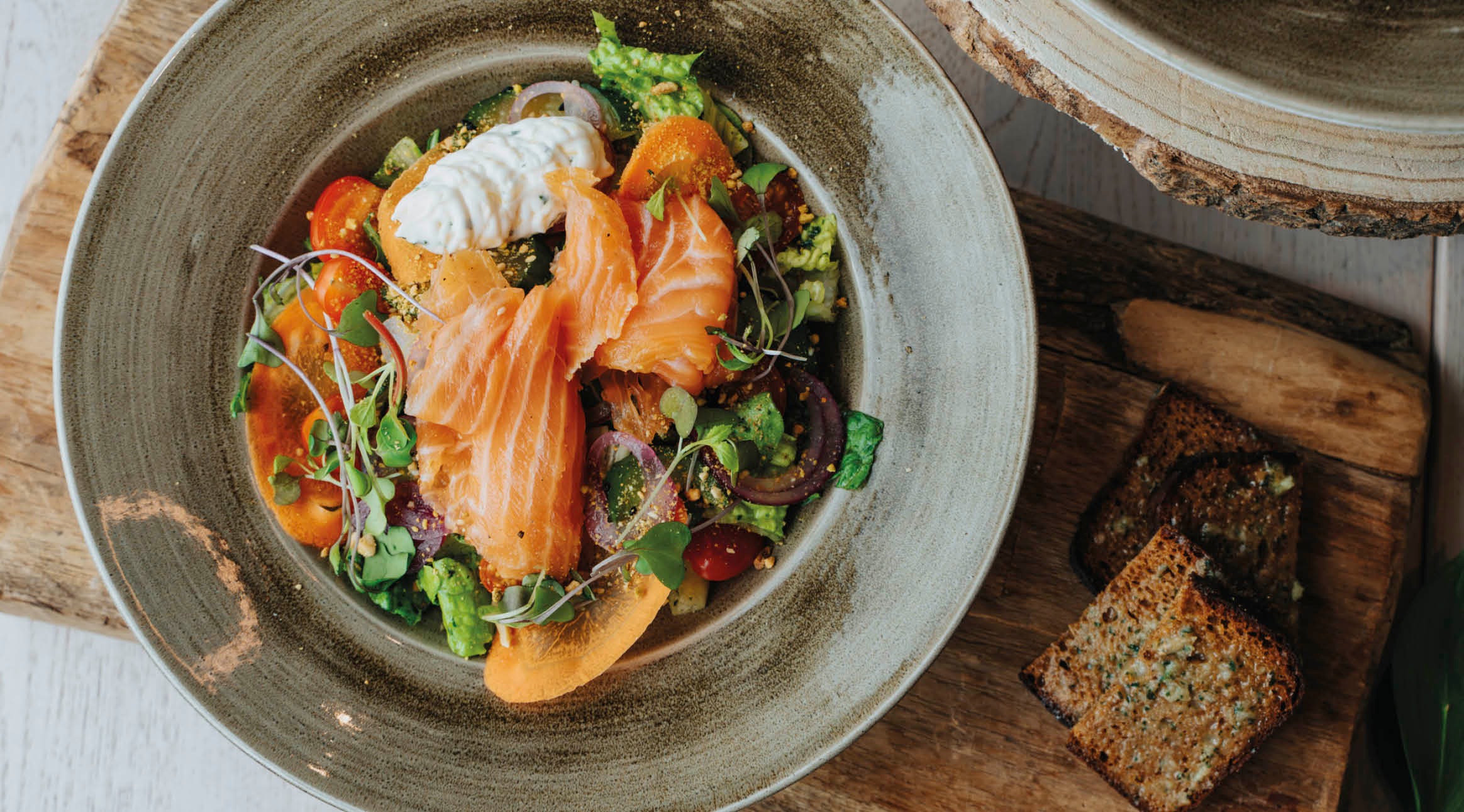 Lohisalaatti keraamisessa kulhossa, sisältää avokaadoa, tomaatteja, versoja ja kermaista kastiketta, tarjoiltuna leivän kanssa puulautasella.