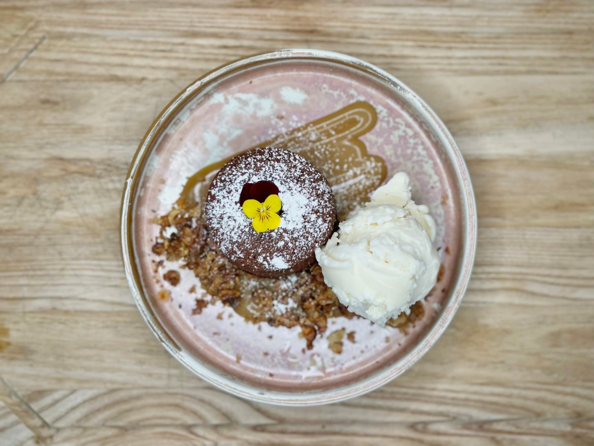 Chocolate cake dusted with powdered sugar, garnished with yellow pansy, served with a scoop of ice cream on a pink plate.