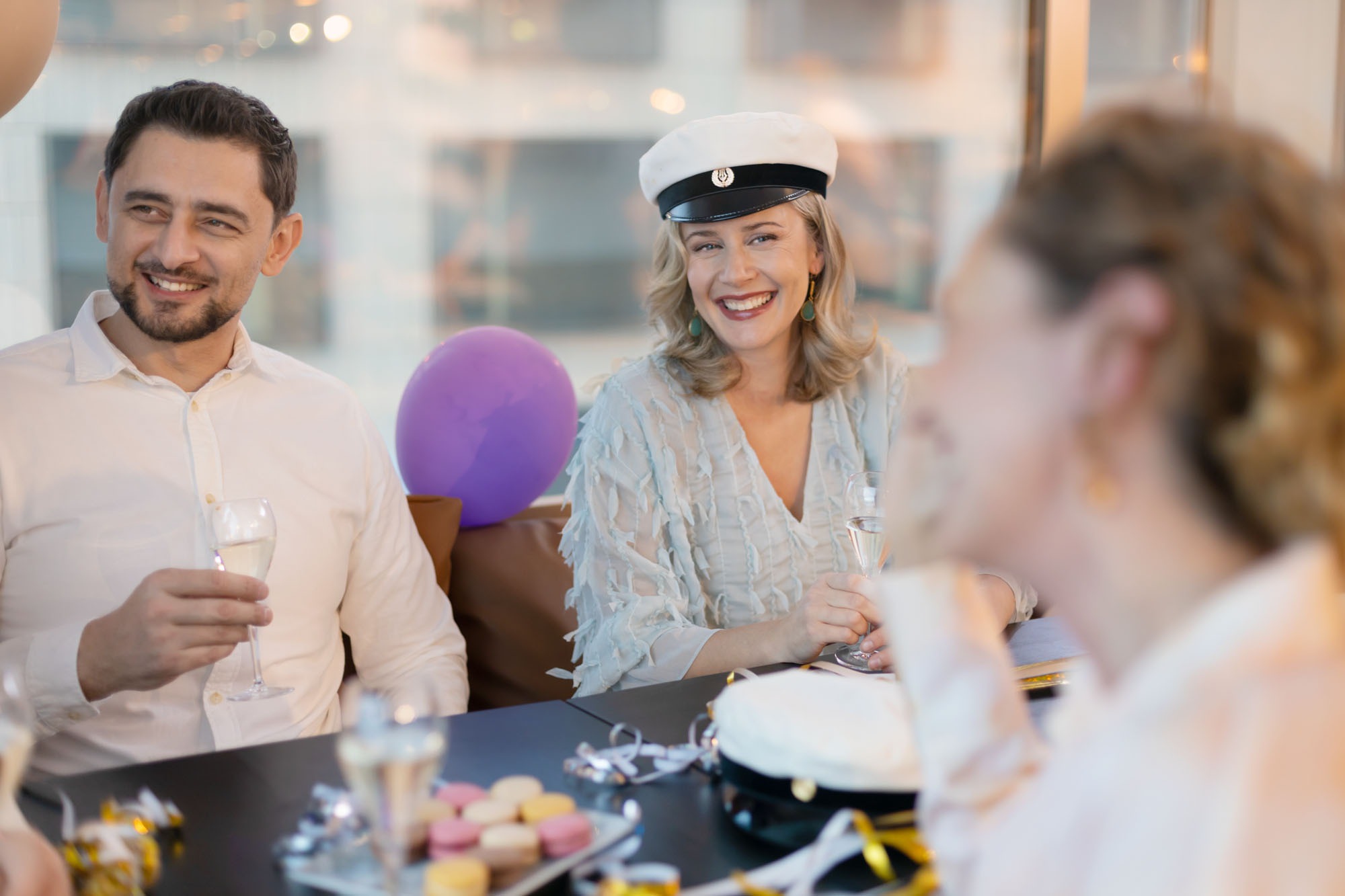 Festive group enjoying champagne at celebration. Person wearing white graduation cap smiles broadly. Purple balloon visible in background.