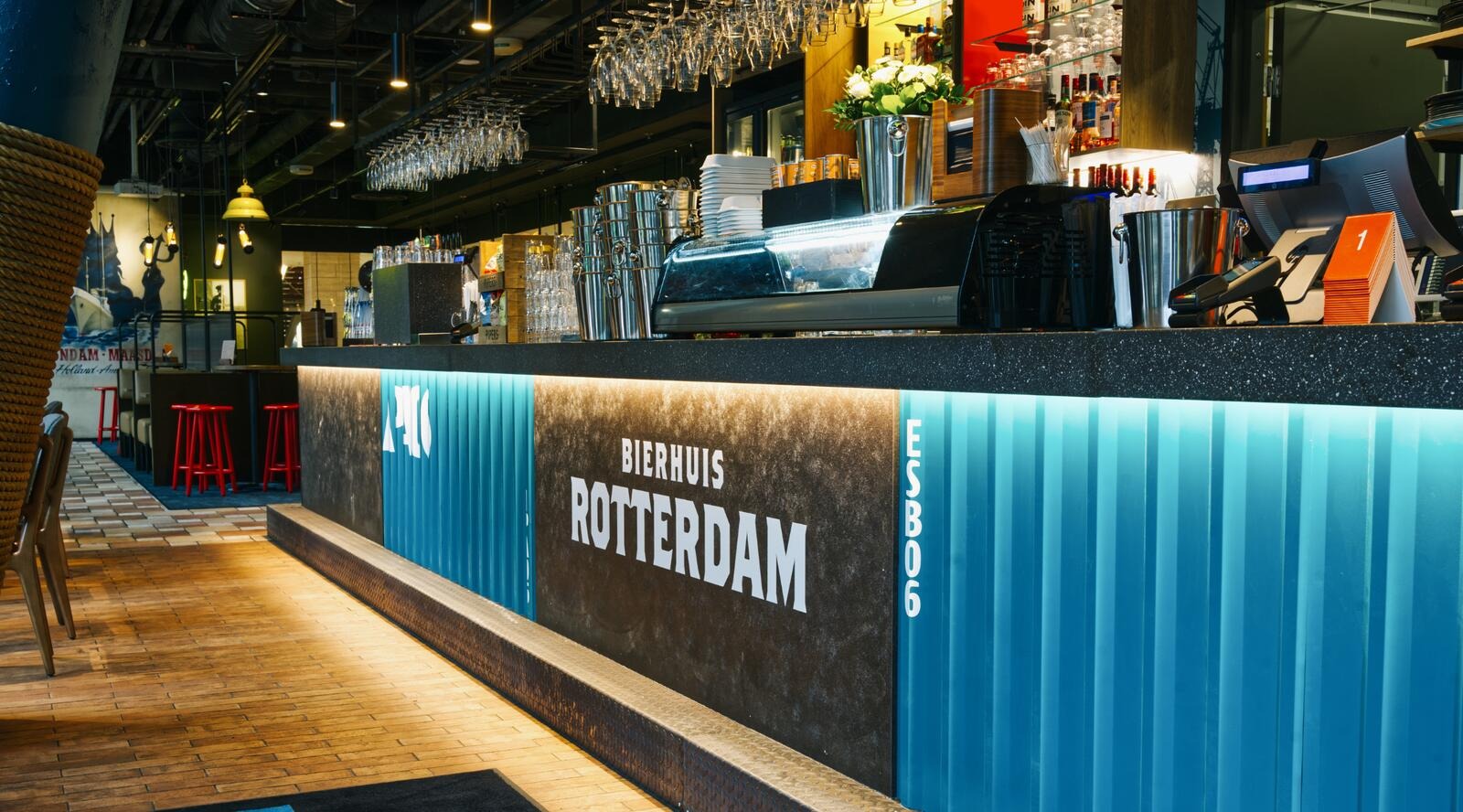 Baari, jossa sininen ja tumma tiski, puinen lattia ja punaisia jakkaroita. Katossa viinilaseja, tiskillä valkoinen kukkakimppu.