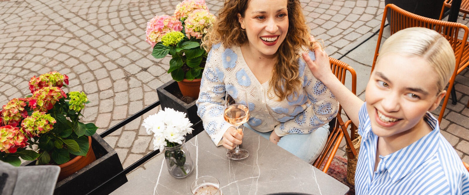 Två personer sitter utomhus vid ett bord på en stenbelagd terrass, blomkrukor bredvid bordet.
