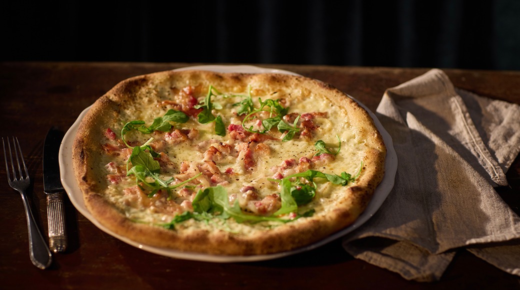 Kergelt röstitud põhjaga pitsa, kaetud juustu, singi ja värskete rukolalehtedega, serveeritud taldrikul puulaual.