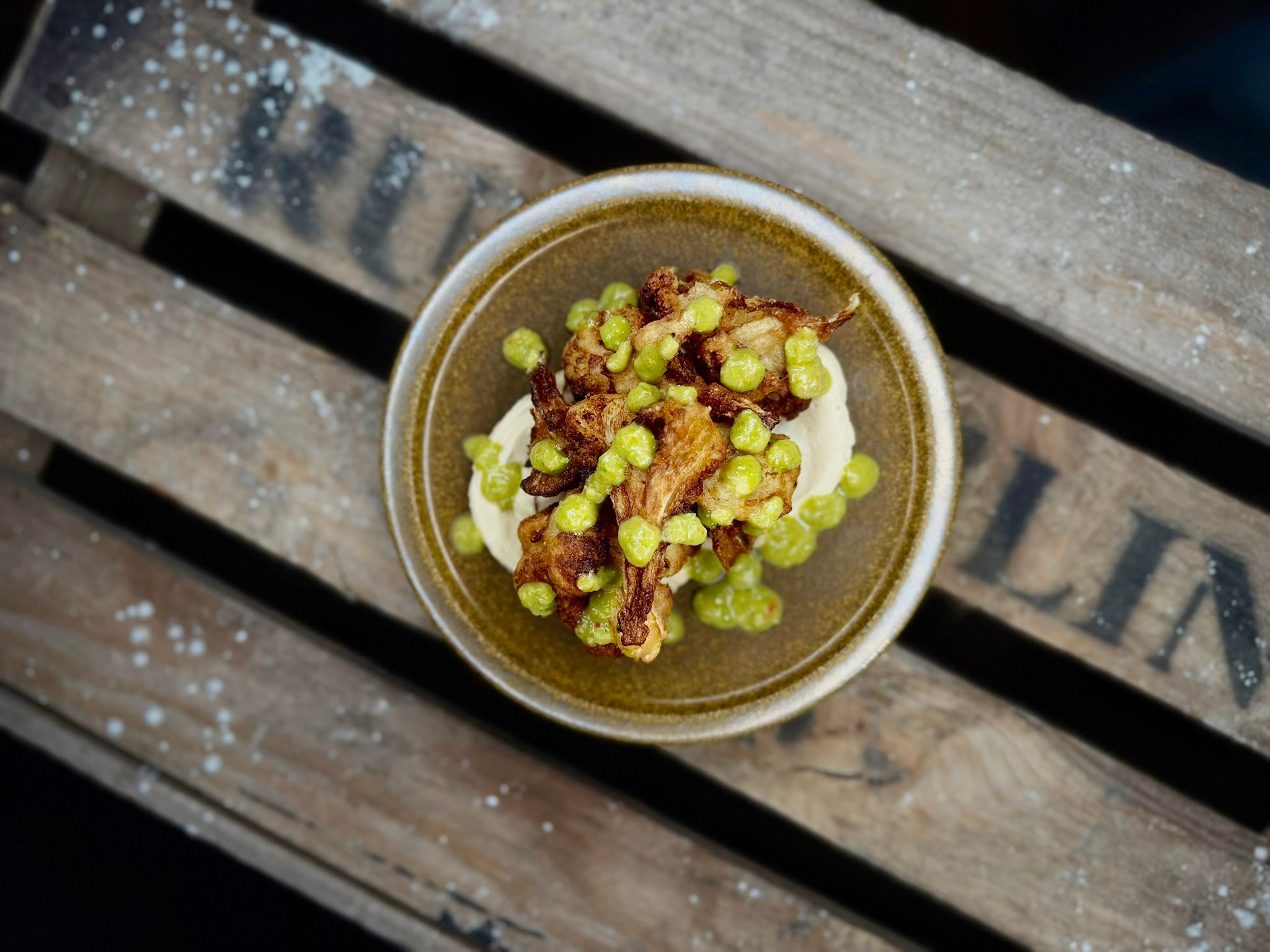 Ceramic bowl containing fried food and green peas on a light sauce, placed on a wooden surface.