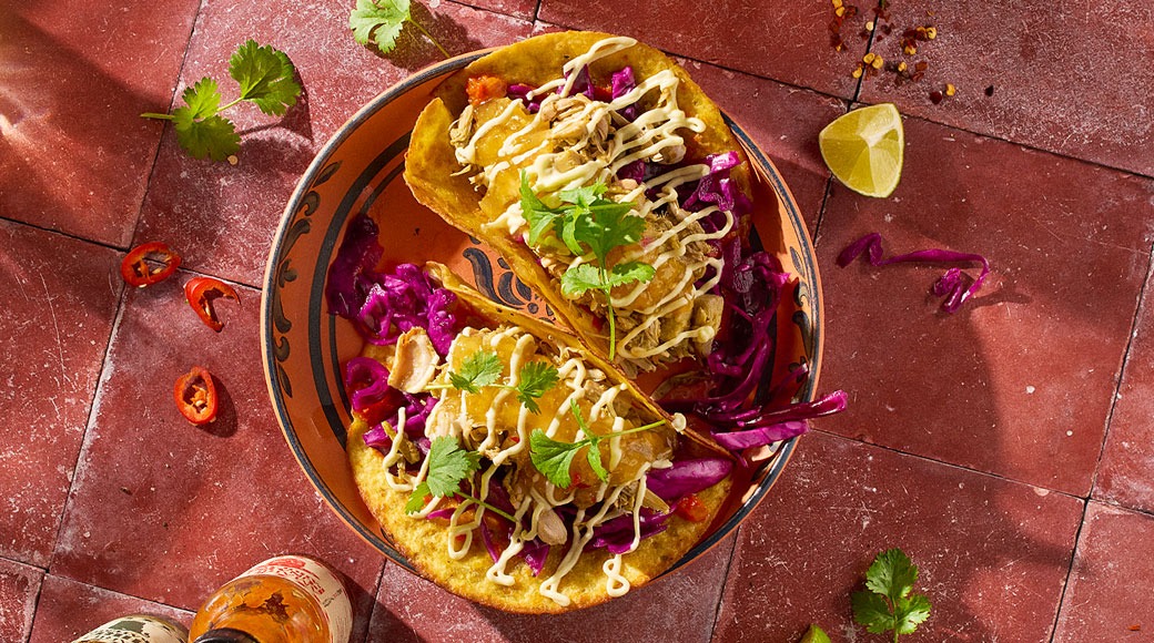 Kaksi tacoa asetettuna koristeelliselle lautaselle punaisella laattalattialla. Täytteenä on pulled-lihaa, ananasta, punakaalia, kastiketta ja korianteria. Vieressä on limetinpuolikas, chilirenkaita ja kuuma kastike.