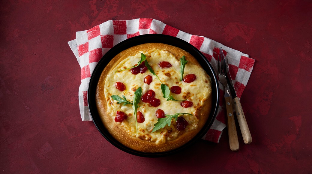 Cheese pizza topped with red berries and rocket in a black pan. A red and white checkered cloth, a knife and a fork lie beside it.