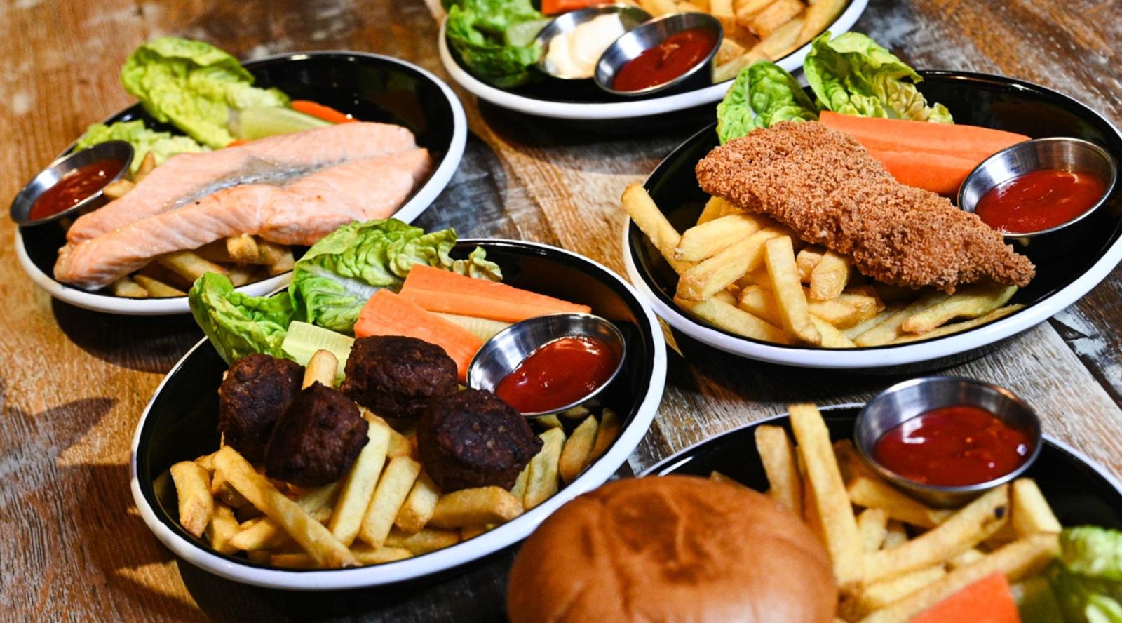 Flera maträtter på restaurang: grillad lax, panerad fisk, köttbullar och hamburgare. Alla serveras med pommes frites, sallad och dippsås.