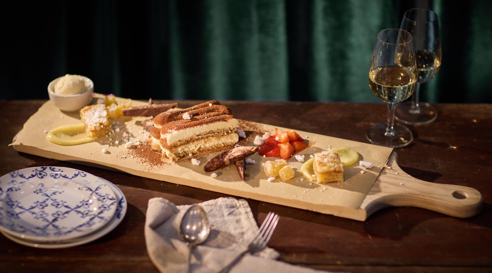 Wooden serving board with tiramisu, fruits and dessert pieces. Alongside ice cream, two wine glasses and a plate.