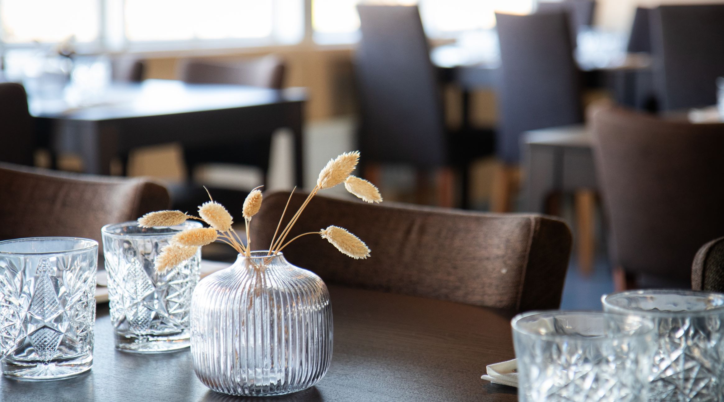 Dried grass stems in a glass vase on a restaurant table, surrounded by crystal glasses. Dark chairs in the background.