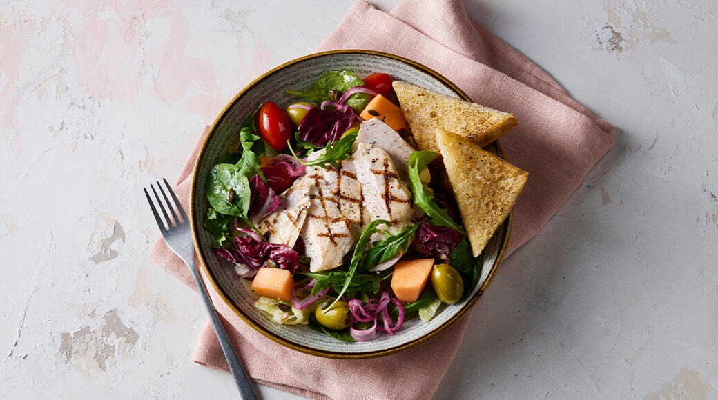 Bowl of grilled chicken salad with tomatoes, melon, olives and red onion, plus two toasted bread triangles. A fork and pink napkin lie beside it.