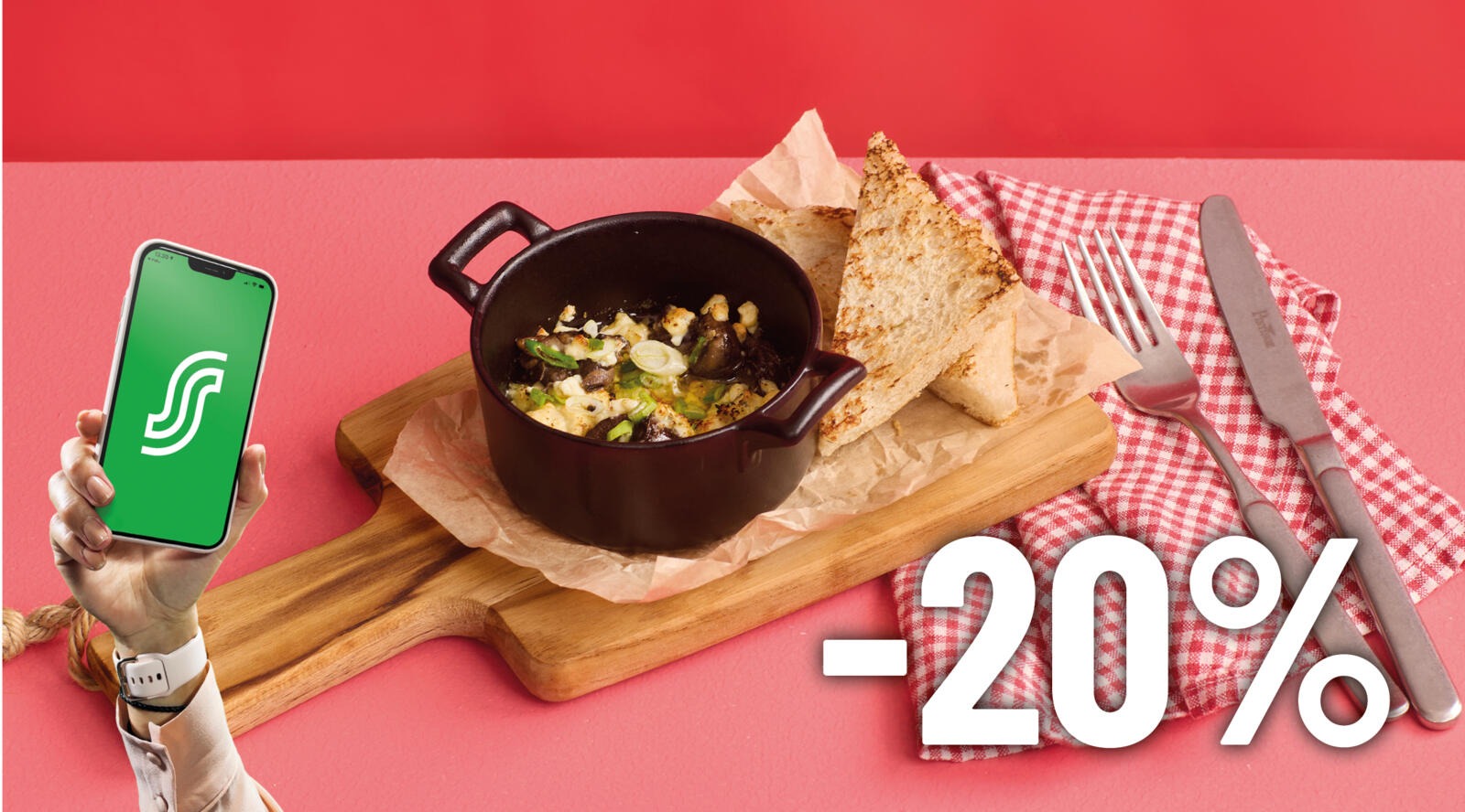 Puhelimessa näkyy vihreä sovellus, vieressä puulaudalla musta kattila, jossa ruokaa, sekä leipää ja ruokailuvälineitä ruutuliinan päällä vaaleanpunaisella alustalla.