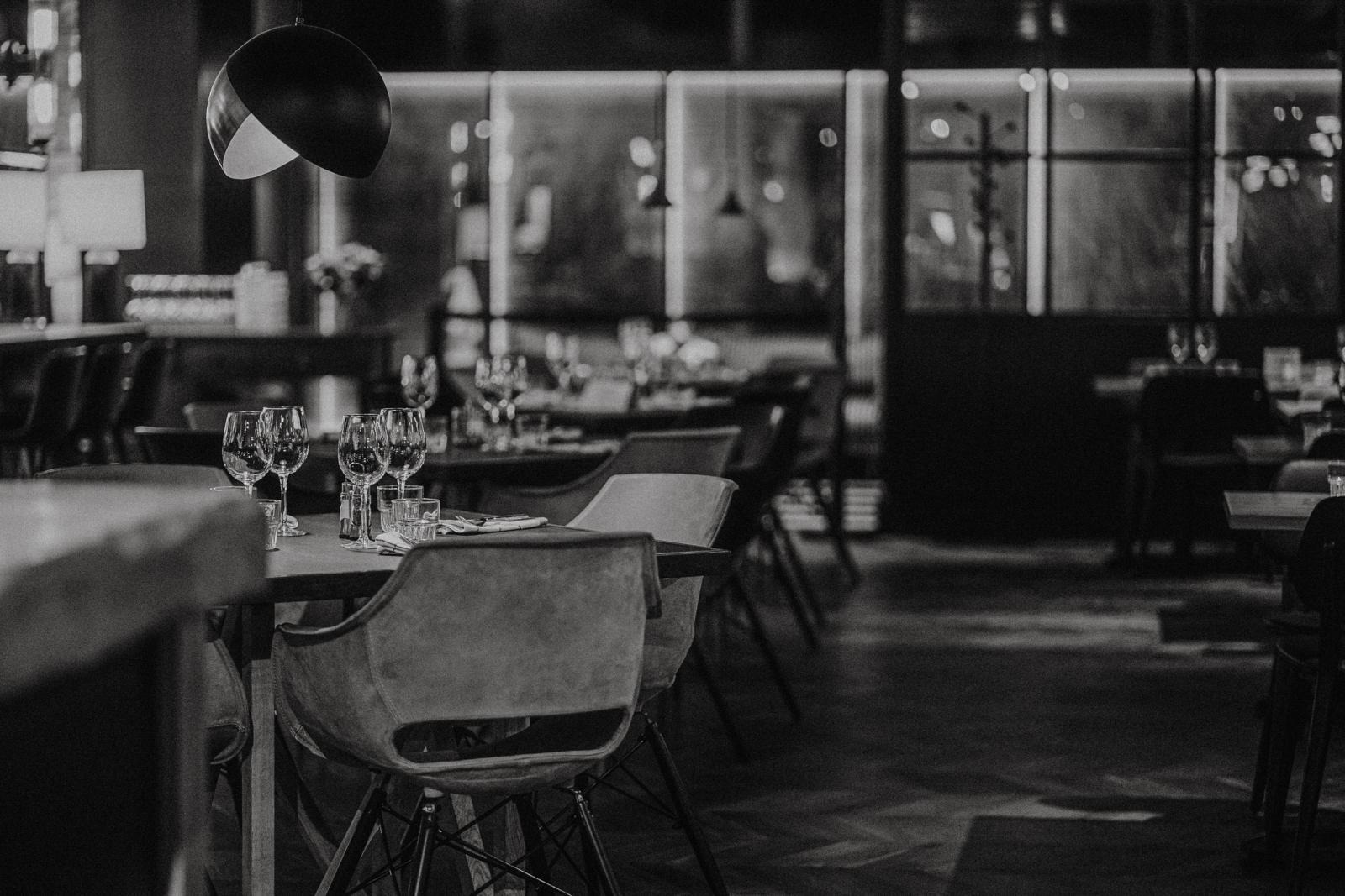 Elegantne restorani interjöör hämaras valguses, laual veiniklaasid ja lauanõud. Avar põrandaplaan modernsete toolide ja rippuvate lampidega.