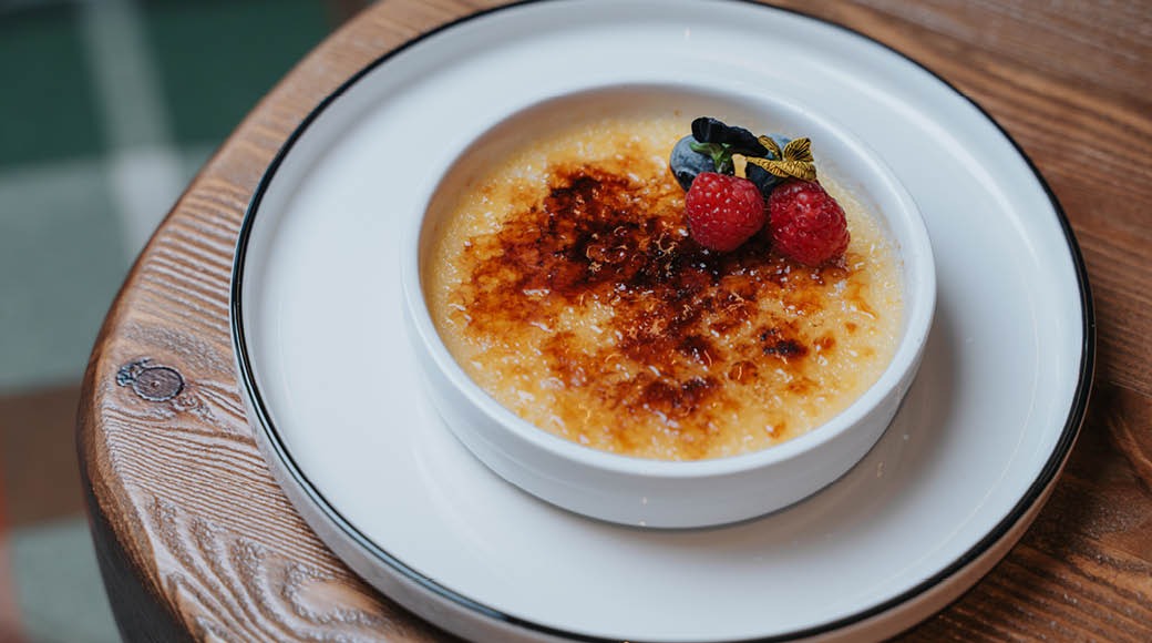Crème brûlée tarjoiltuna valkoisessa kulhossa, päällä vadelmia ja mustikoita, puisella pöydällä.