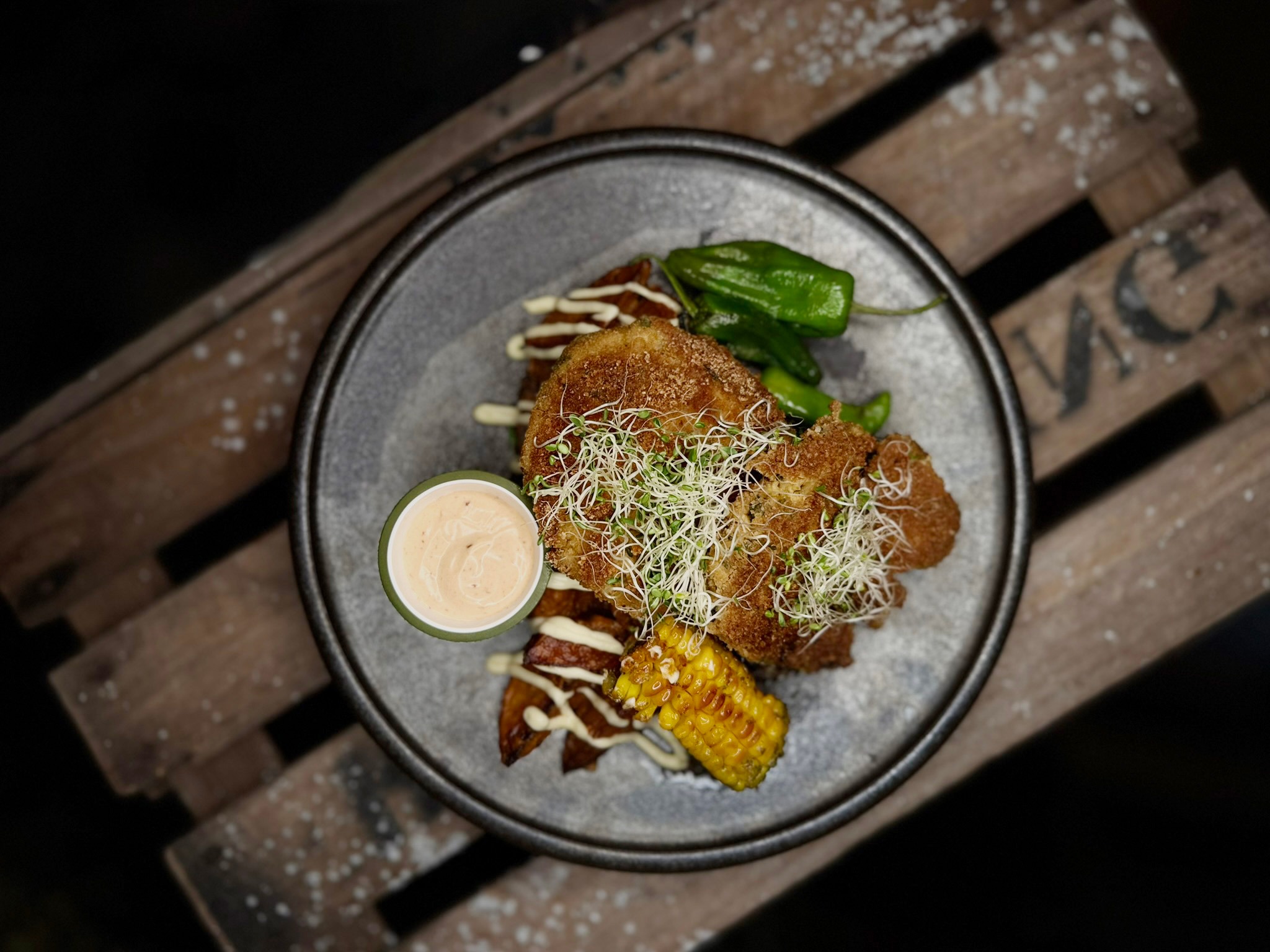 Gray plate with breaded food, microgreens, green chili pepper, corn, and dipping sauce on a wooden surface.