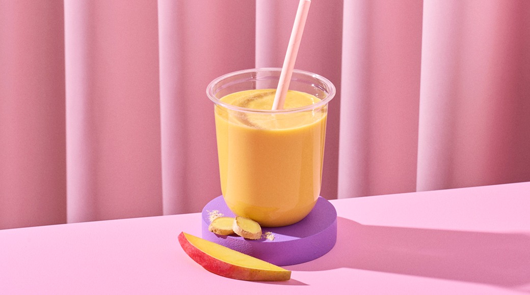 Keltainen mangosmoothie kirkkaassa muovimukissa pillillä violetin alustan päällä. Edessä mangolohko ja inkiväärin viipaleita vaaleanpunaista taustaa vasten.