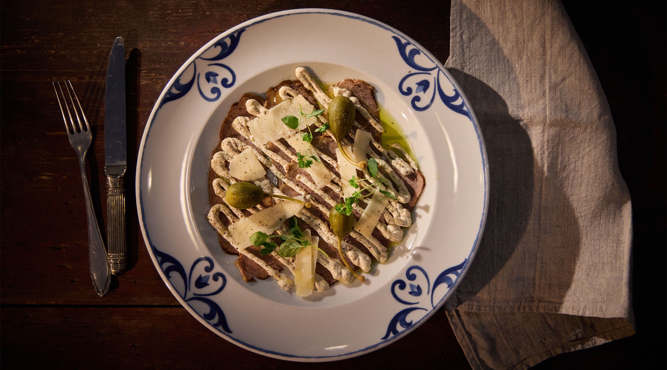 Annos pihvilihaa valkoisella sinireunakoristelulla lautasella, päällä juustolastuja, oliiveja, kastiketta ja tuoreita yrttejä.