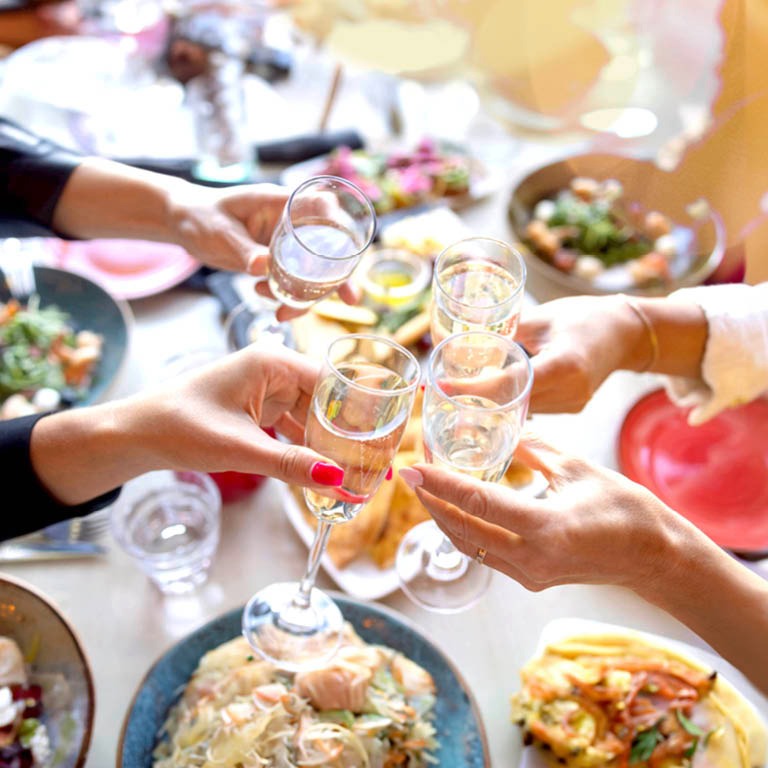 Neljä kättä kohottaa laseja maljaksi juhla-aterian ääressä, pöydällä näkyy erilaisia ruoka-annoksia lautasilla.