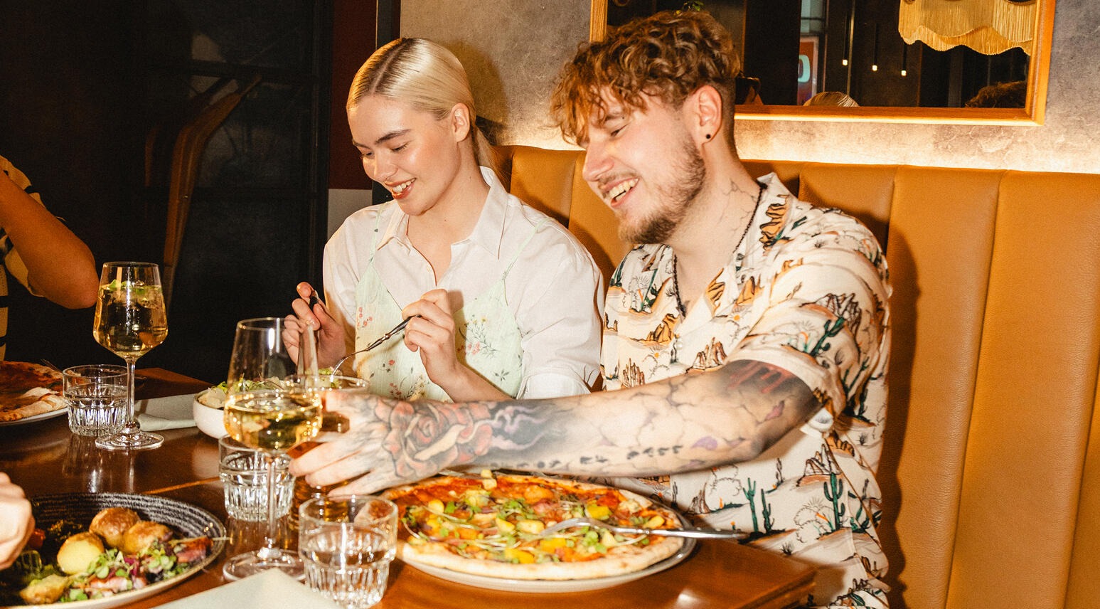 Kaksi henkilöä nauttii ruokaa ravintolan pehmusteisessa penkkipöydässä. Ruokavalikoimaan kuuluu pizza ja salaatteja.