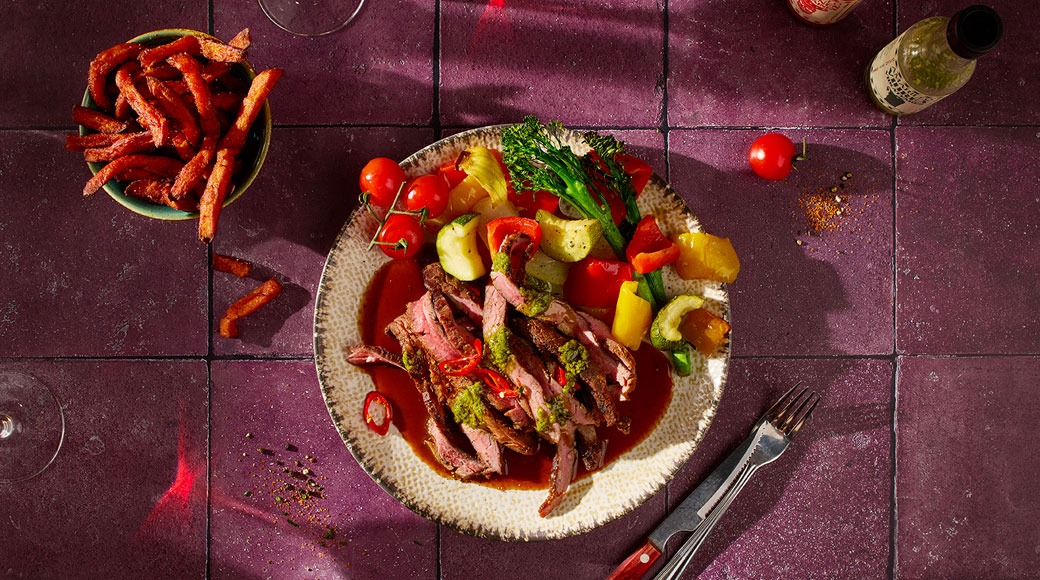 Viilutatud grillitud liha punases kastmes, tomatite, paprika, suvikõrvitsa ja brokoliga taldrikul. Kõrval kauss bataadifriikartuleid ja söögiriistad lillakatel plaatidel.