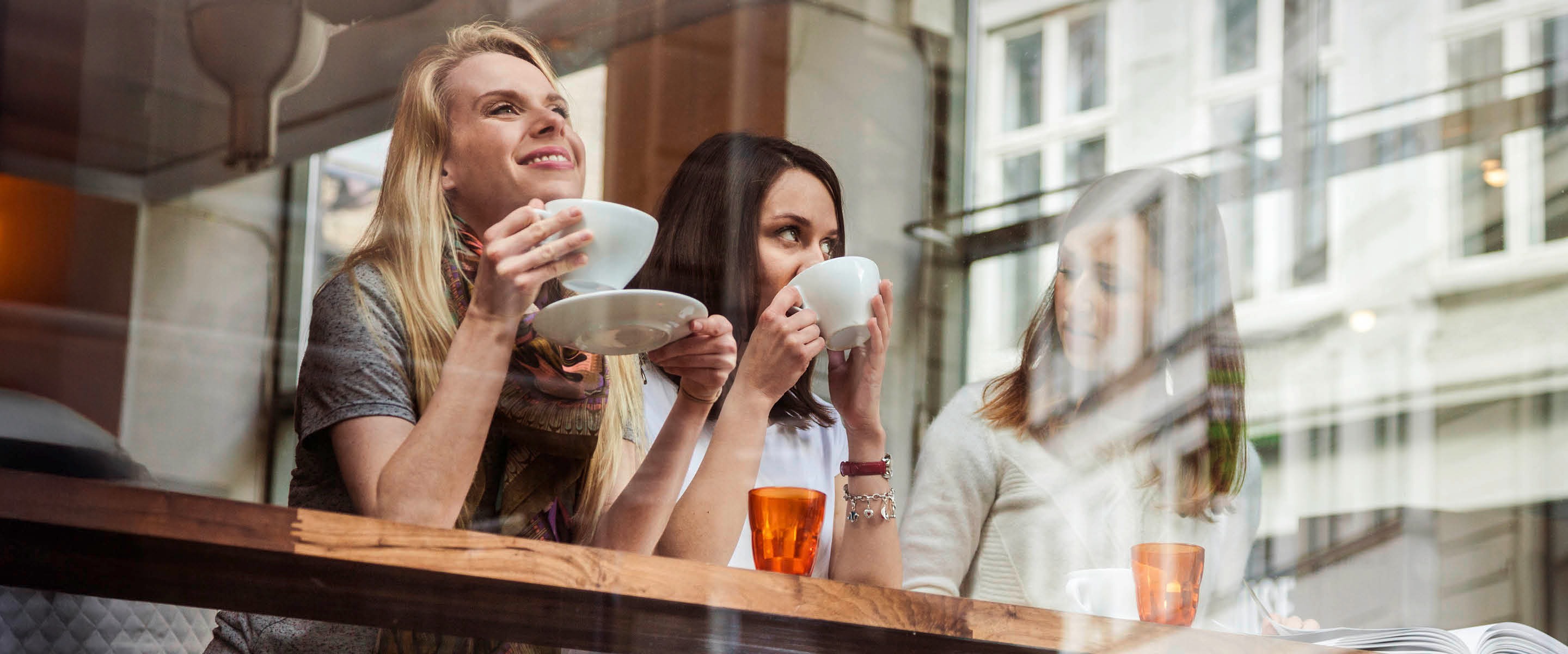 Kolme henkilöä nauttimassa kahvia kahvilassa ison ikkunan ääressä, pitäen valkoisia kuppeja käsissään. Pöydällä on oransseja juomalaseja.