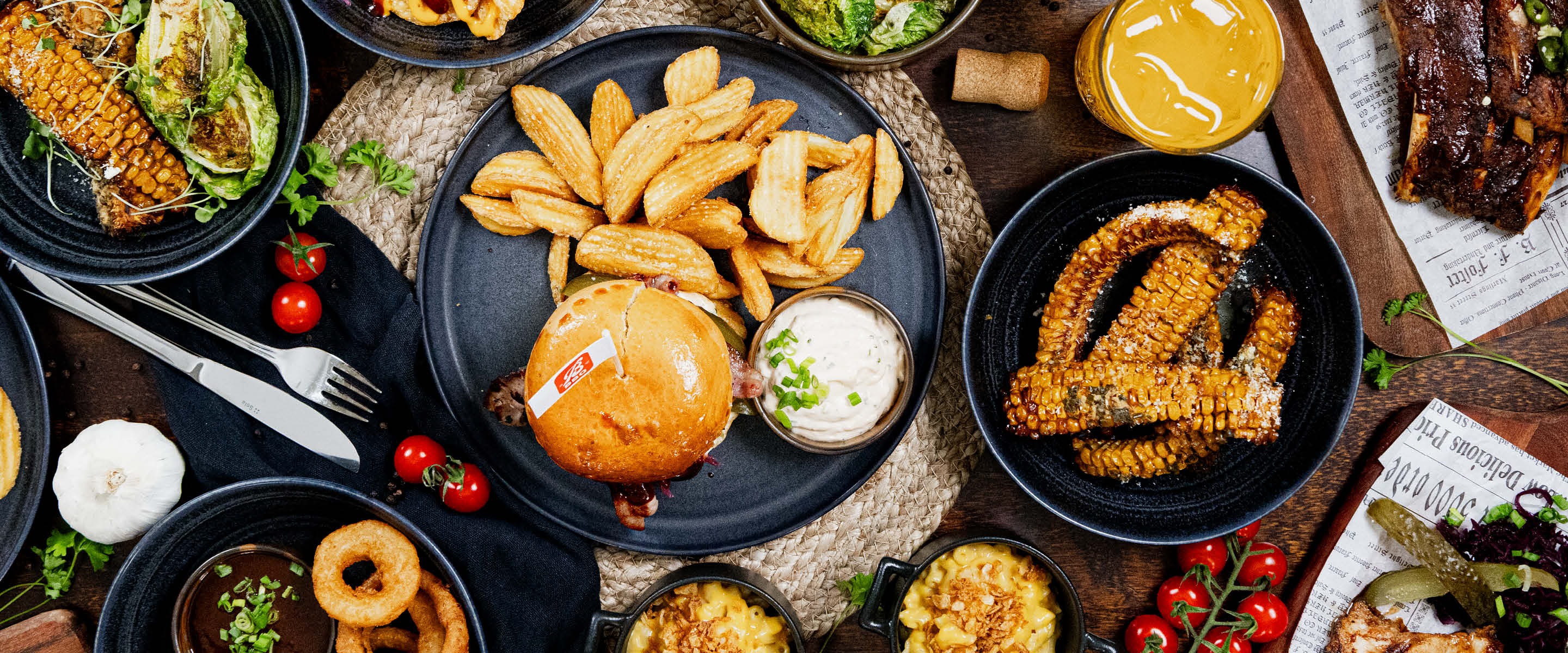 Abundant spread of restaurant food: burger with fries, grilled corn, ribs, vegetables and dipping sauces on black plates arranged on wooden surface.