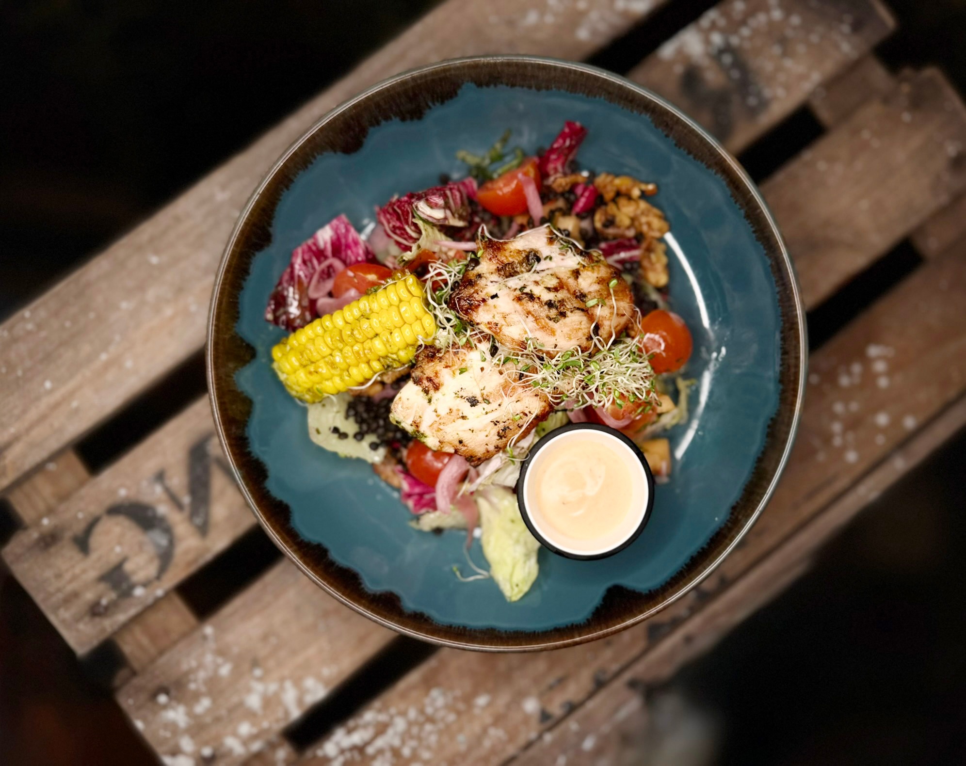 Grilled meat with corn, tomatoes, salad and dipping sauce on a blue plate atop a wooden surface viewed from above.
