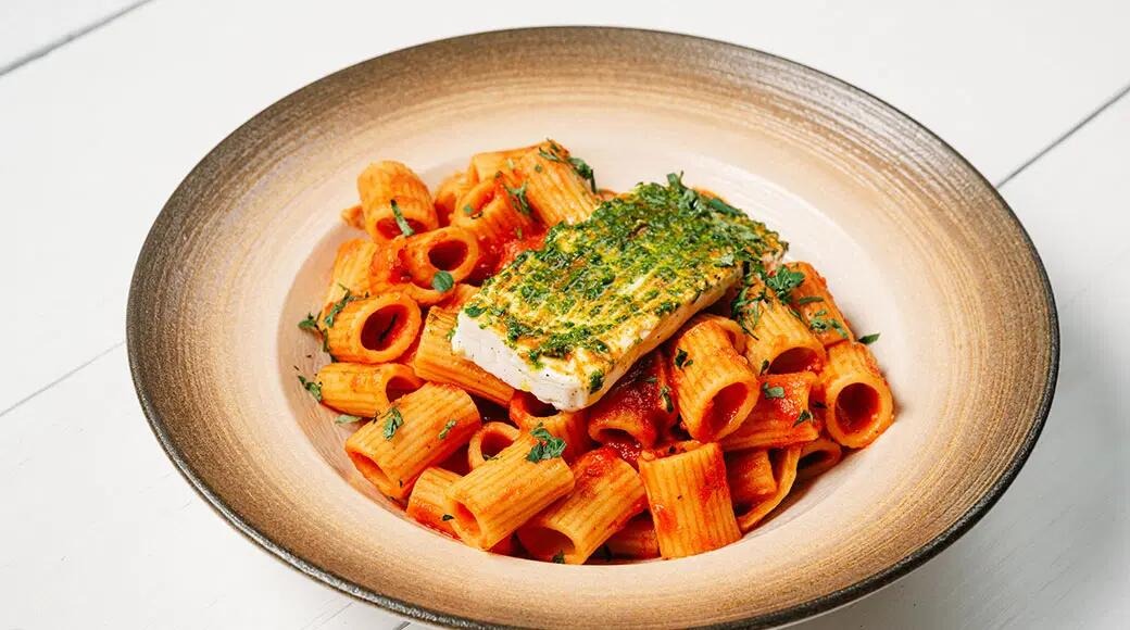 Rigatoni pasta tomatikastmes, peal ürtidega kaetud fetajuustu tükk. Roog on serveeritud laias pruunikas kausis.