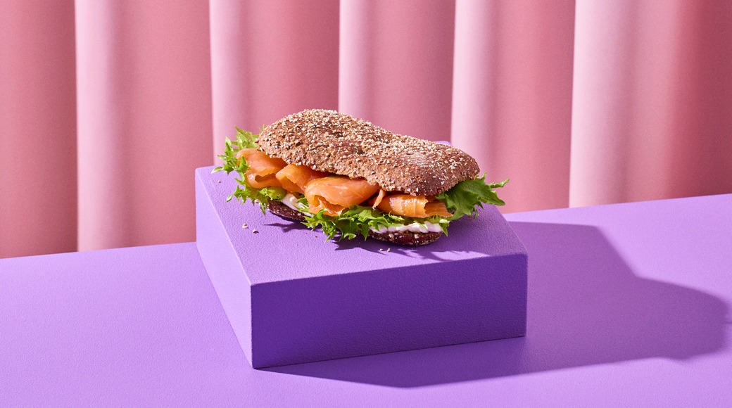 Täysjyväleipä, jossa on graavilohta, salaattia ja tuorejuustoa. Voileipä on violetin korokkeen päällä vaaleanpunaista verhoa vasten.