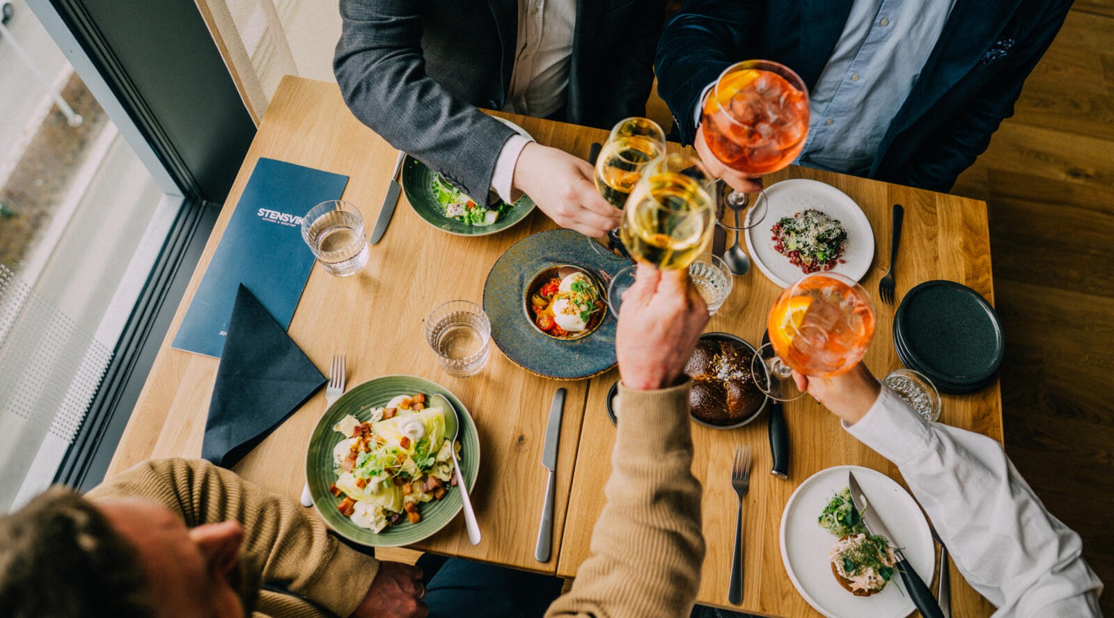 Händer som skålar med vin och cocktailglas över ett middagsbord. Träbord fullt med färgglada rätter och sallader.