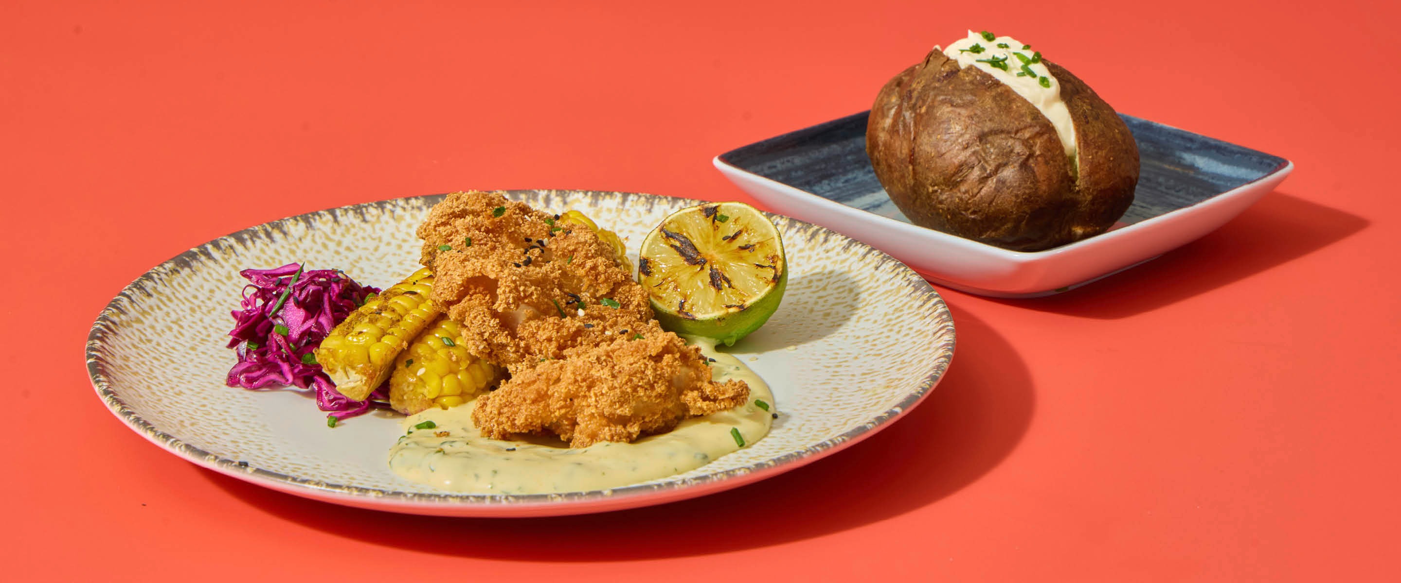 Panerad mat, majs, rödkål och en limeklyfta på en tallrik, bredvid en bakad potatis med fyllning på ett litet fat. Korallröd bakgrund.