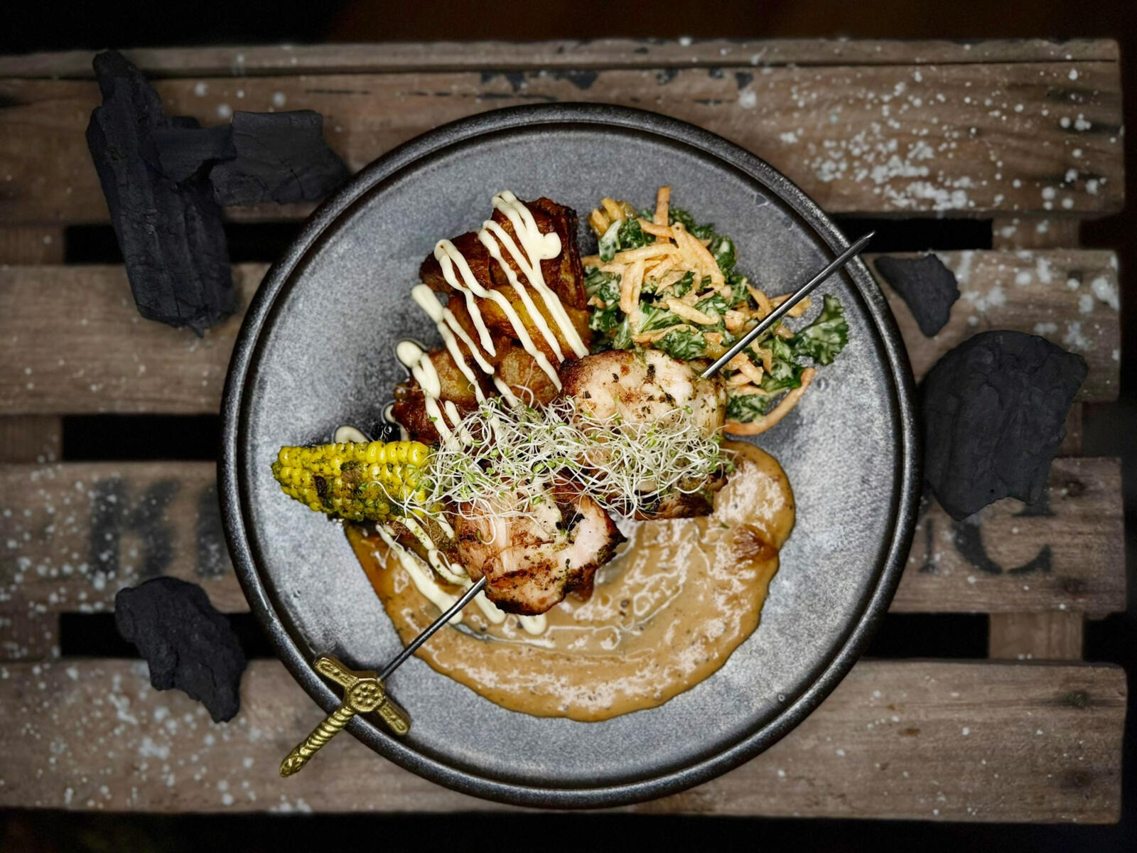 A gray plate featuring grilled meat, sauce, microgreens, yellow corn, and salad, placed on a wooden surface with decorative charcoal pieces.