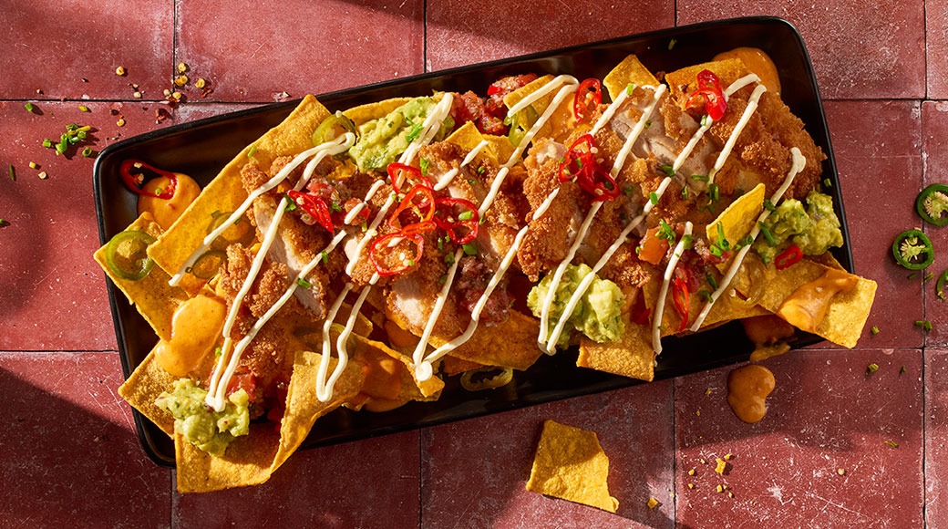 Mustalla tarjottimella nachoja, joiden päällä on rapeaa kanaa, guacamolea, salsaa, chiliviipaleita ja valkoista kastiketta.
