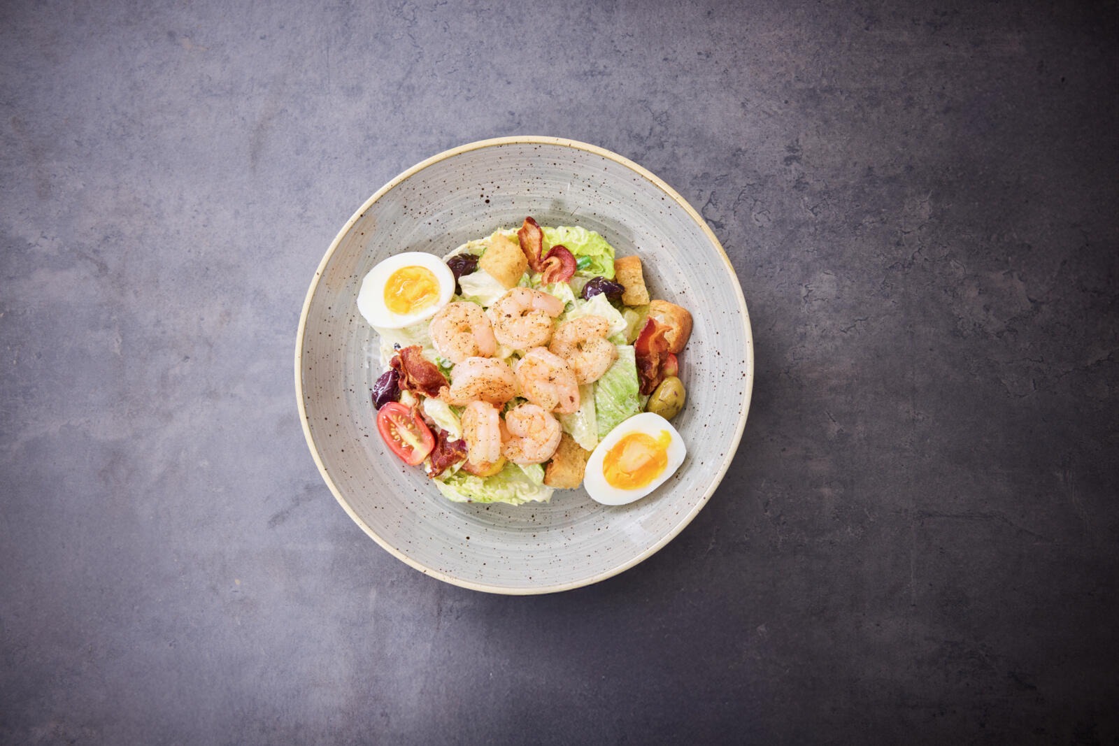 Katkarapusalaatti keraamisessa kulhossa, jossa on kananmunan puolikkaita, tomaatteja ja salaattia harmaalla taustalla.