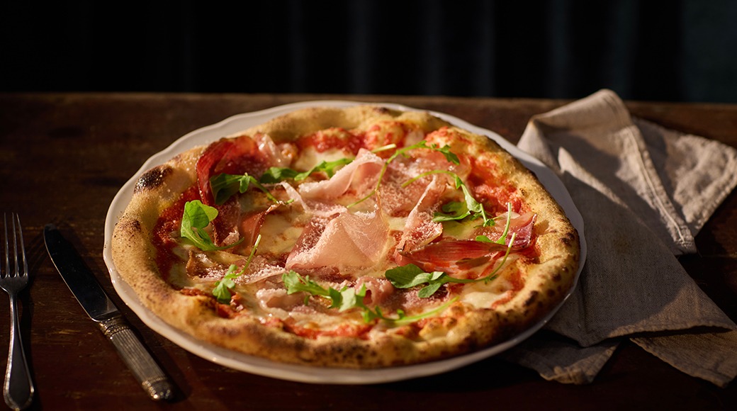 Käsitööpitsa prosciutto, tomatikastme ja värske rukolaga, serveeritud valgel taldrikul puulaual.