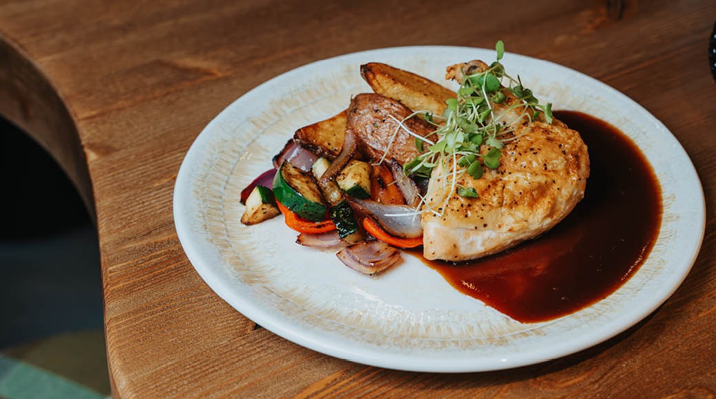 Kana-annos valkoisella lautasella, jossa paistettua rintafileetä, paahtuneita juureksia ja kasviksia sekä tummaa kastiketta.