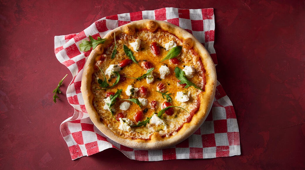 Pizza topped with melted cheese, cherry tomatoes, fresh cheese and arugula. The pizza is on a red and white checkered cloth on a red background.
