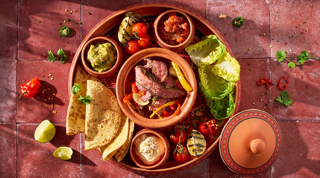 Terrakottalautasella grillattua lihaa, tortillat, guacamole, salsa, smetana, salaattia ja kirsikkatomaatteja punaisella laatalla.