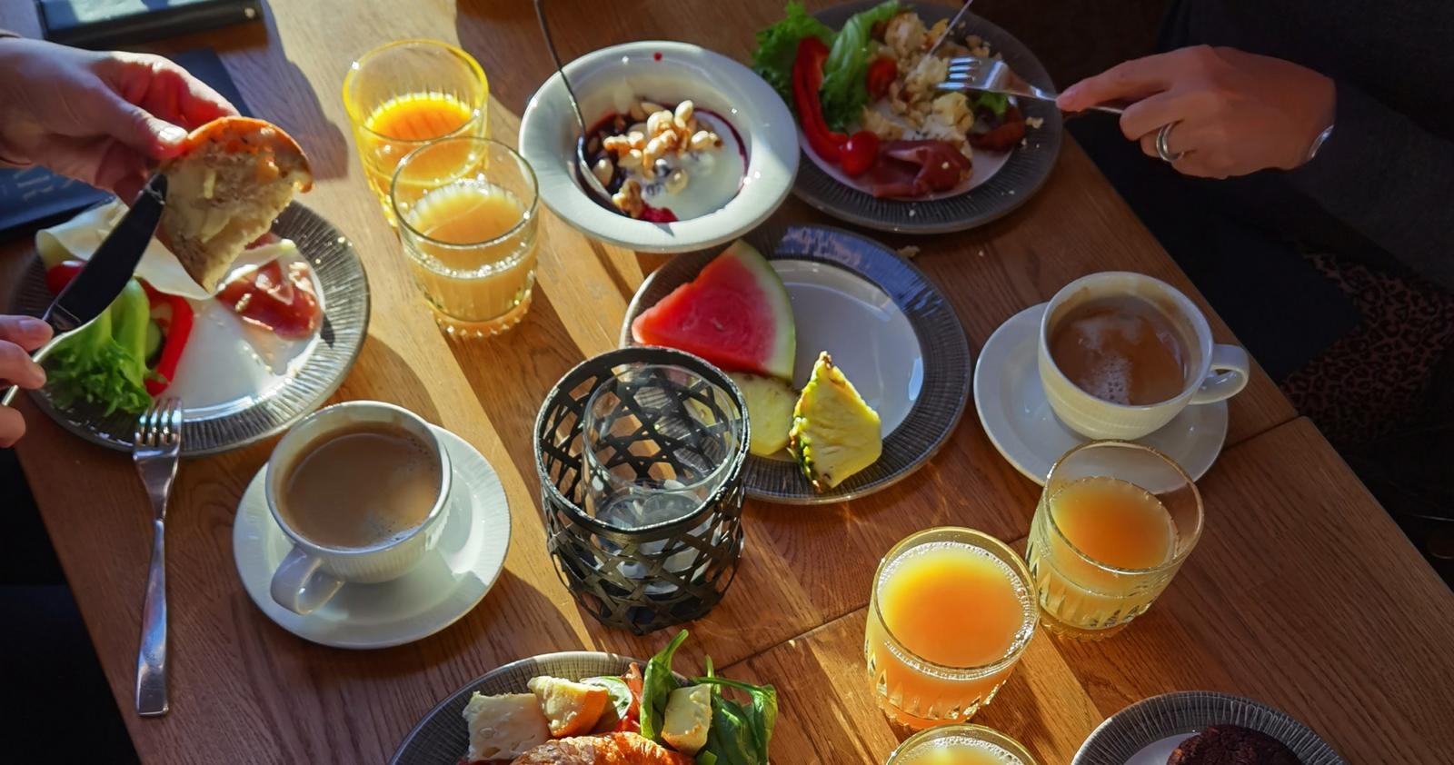 Hommikusöögilaud täis toitu, sealhulgas puuviljatükid, kohv, mahl ja leib. Näha on söövaid käsi.