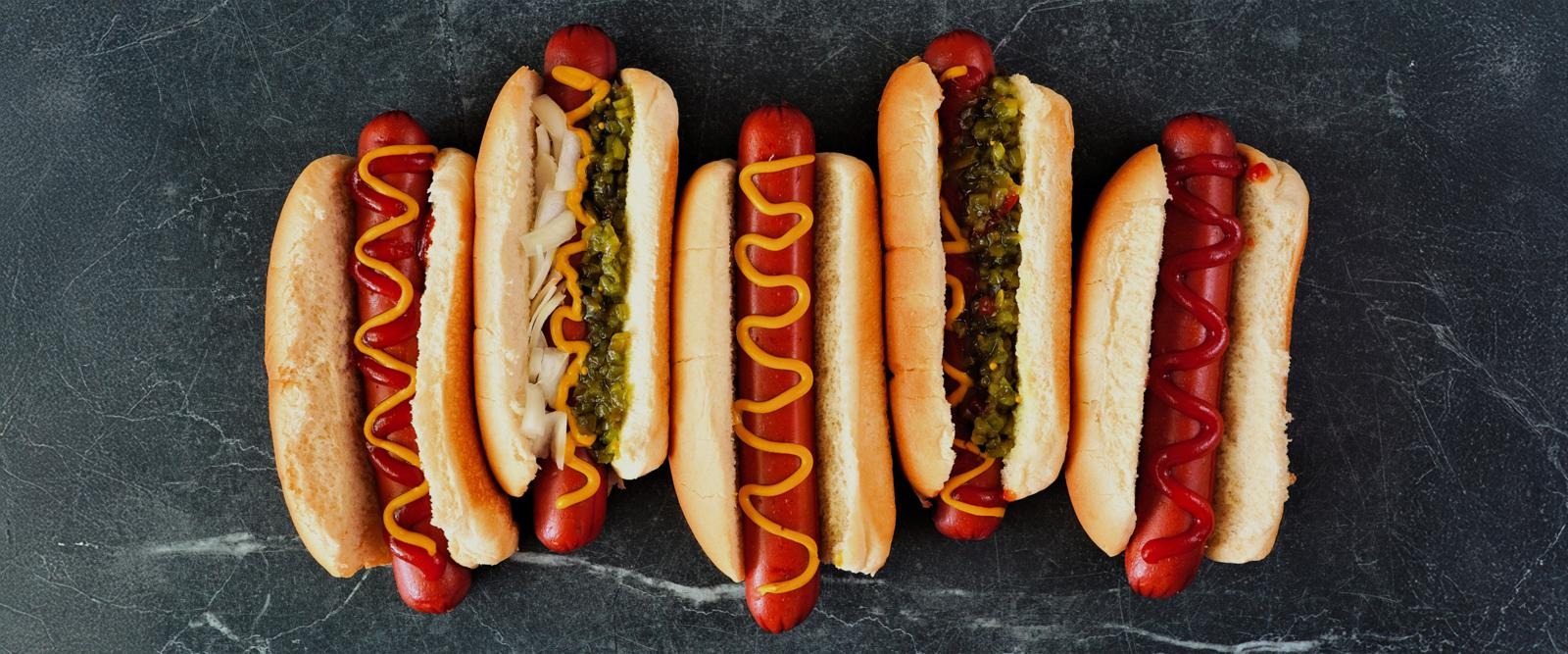 Viisi erilaista hot dogia rivissä tummalla kivipinnalla - perinteisiä sinapilla ja ketsupilla sekä raikkailla lisukkeilla.