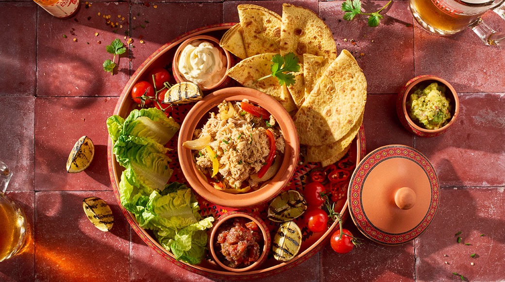 Yläkuva meksikolaisesta ruoka-annoksesta punaisella tarjottimella: tortillakolmioita, suikaloitua lihaa, salaattia, grillattuja limetin puolikkaita, salsaa, guacamolea ja smetanaa.