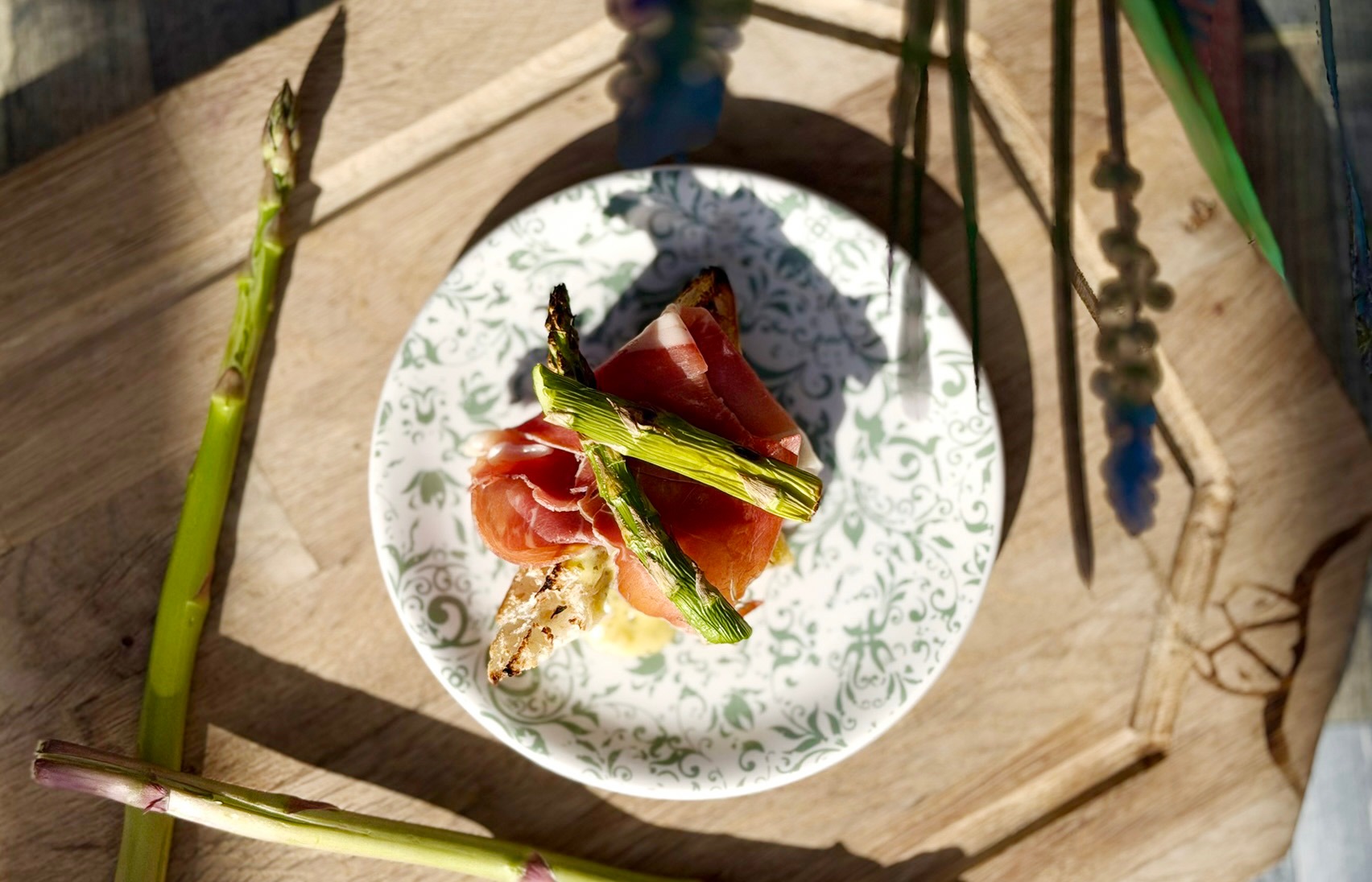 Prosciutto ham and grilled asparagus on a decorative plate on wooden surface. Fresh asparagus stalks surrounding the dish.
