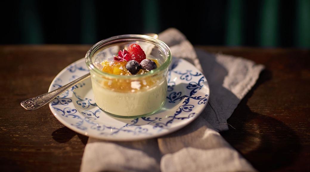 Lasipurkissa tarjoiltu pannacotta, jonka päällä on marjoja ja hedelmäsosetta. Annos on koristellisella lautasella pöytäliinalla.