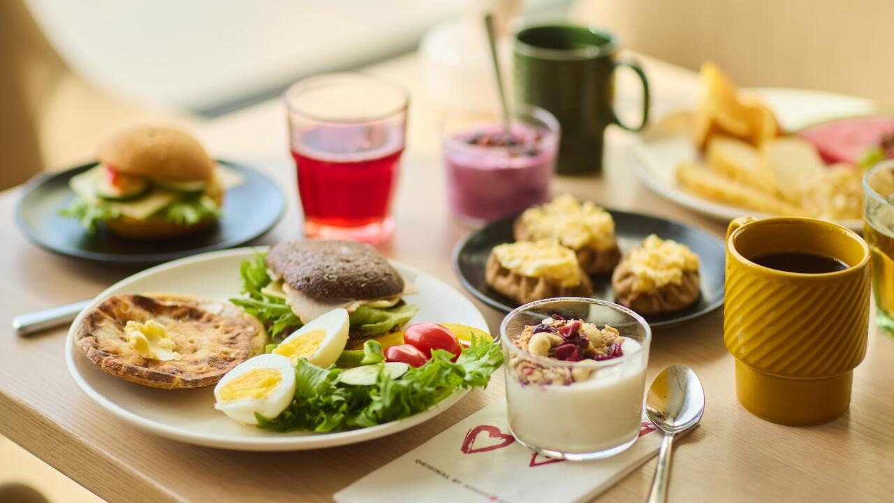 Aamiaispöytä täynnä erilaisia ruokia: sämpylöitä, kananmunia, salaattia, jälkiruokaa ja juomia värikkäissä astioissa puisella pöydällä.