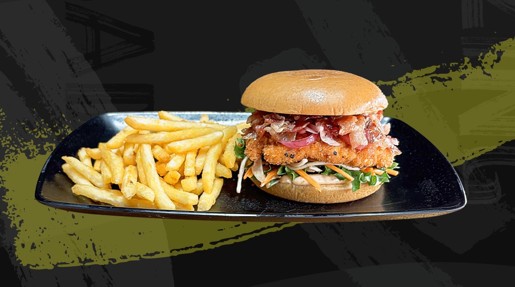 Burger with crispy chicken patty, vegetables and bacon, served with french fries on a black plate.