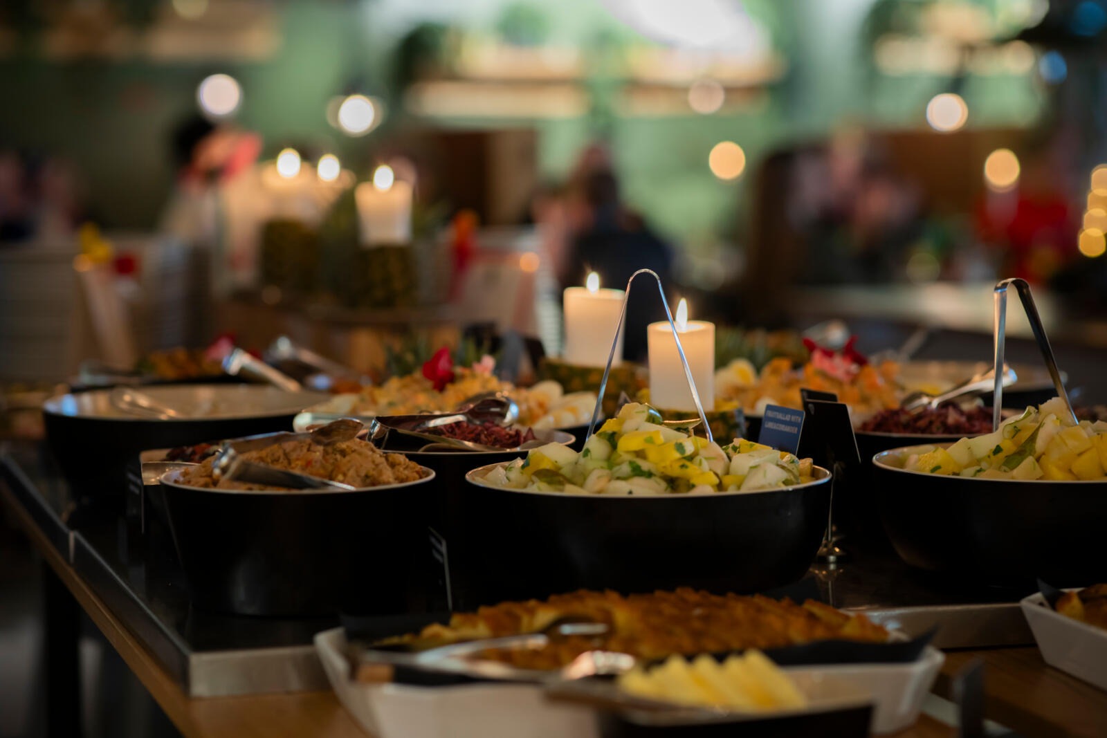 Buffet table filled with various dishes in serving bowls with candlelight, people blurred in background.