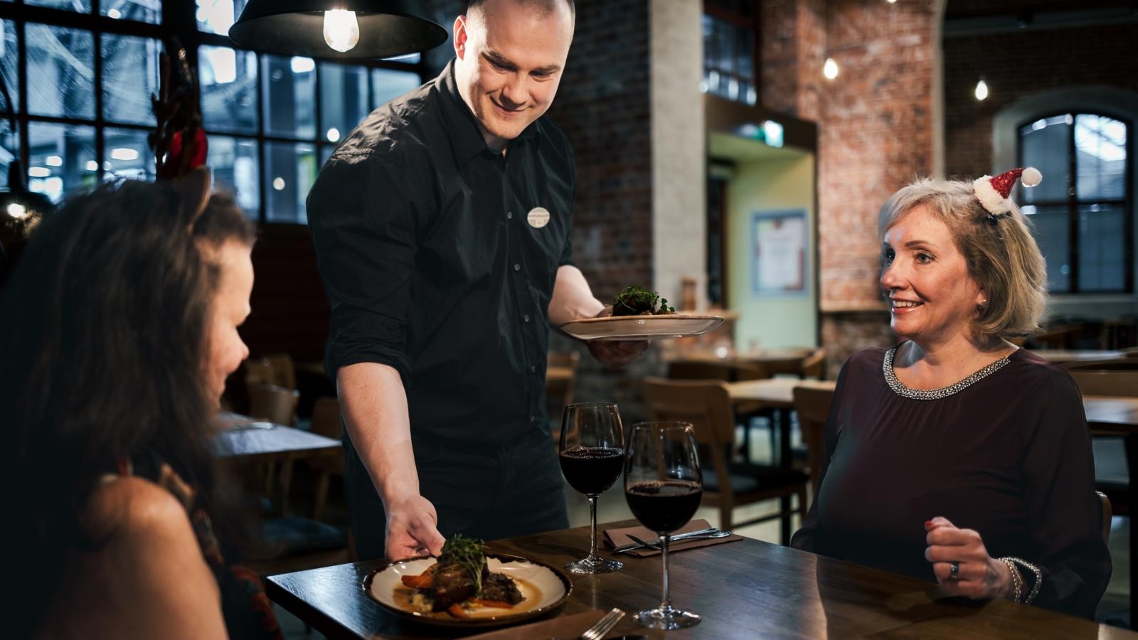 Server in black shirt presenting a plated meal to diners enjoying red wine in a restaurant with exposed brick walls.