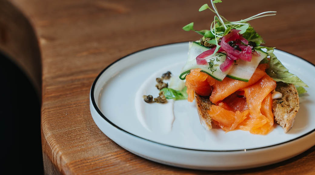 Annos lohivoileipää valkoisella lautasella, koristeena tuoreita kasviksia, punasipulia ja versoja puisella pöydällä.