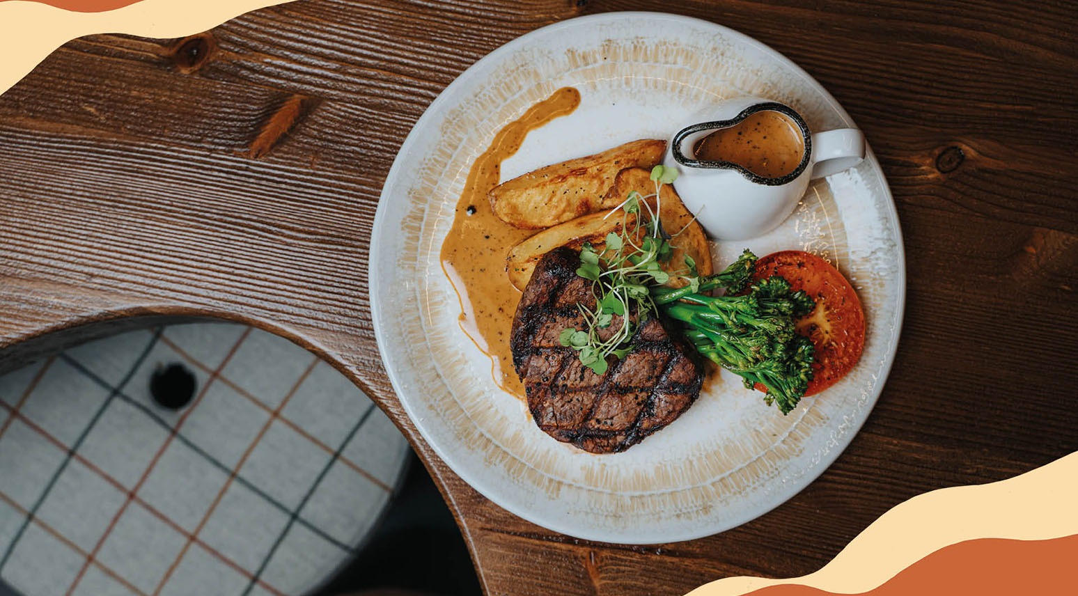 Pihviateria valkoisella lautasella puisella pöydällä, sisältäen grillatun pihvin, parsakaalia, perunoita ja kastiketta pienessä kannussa.