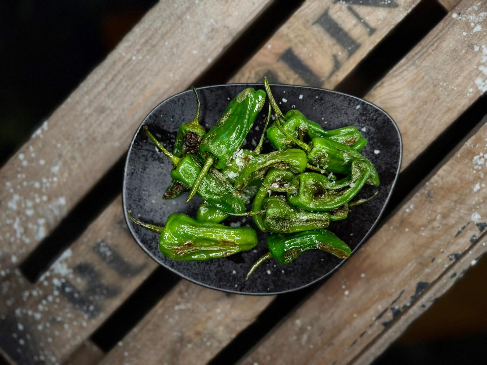Roasted green peppers or padrón peppers on a black plate sitting on a wooden surface.